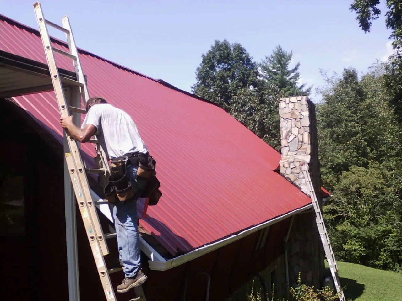 Roofer on a ladder beside a red metal roof and stone chimney — Soffit & Fascia Repair by Roofing Brothers in Valley Head